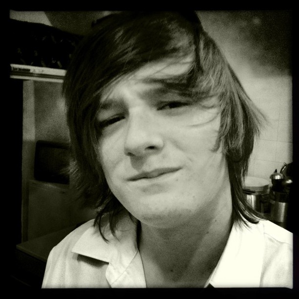 A black and white photograph of a young man with long dark hair styled in a 1970s fashion, wearing a white collared shirt.