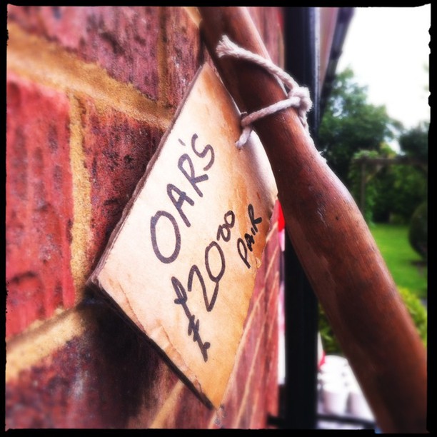 A handwritten price tag reading Oaks +1200 per. is attached to what appears to be a wooden post or tree, with green foliage visible in the background.