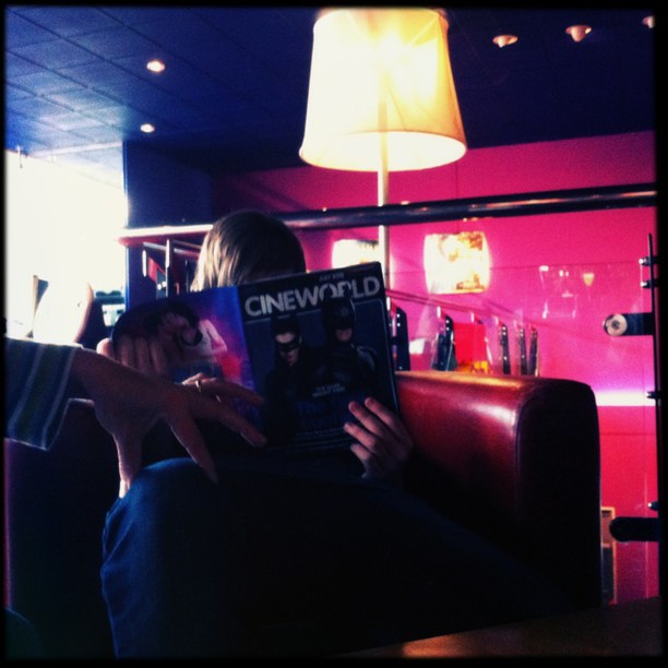 A person holds a magazine covering their face inside a cinema, with pink and blue neon lighting visible in the background.