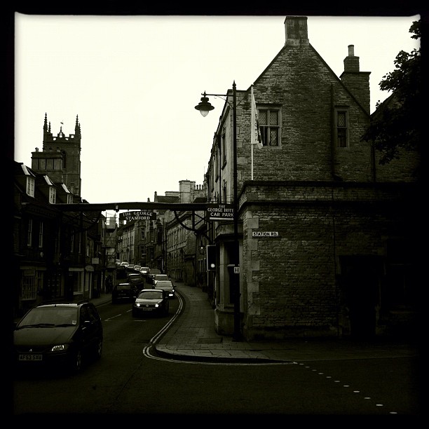 A historic British stone street lined with period buildings, parked cars, and a church tower visible in the distance.