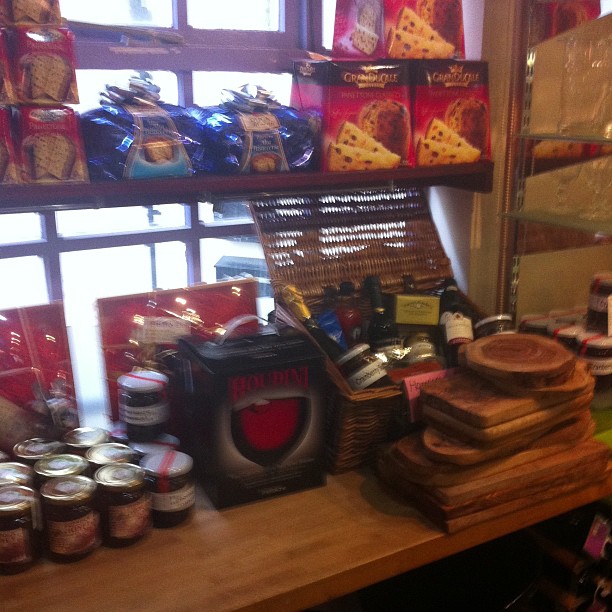 A shop window display featuring packaged biscuits on upper shelves, a red coffee maker, wooden chopping boards, jars, and a wicker basket on the counter below.