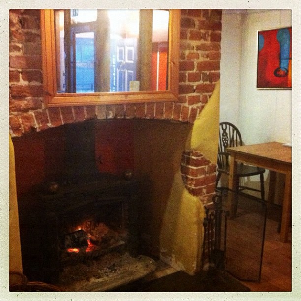 A fireplace with glowing embers burns in a brick alcove. A wooden table and chair are visible to the right in the room.