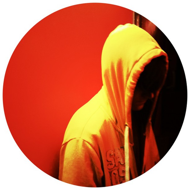 A person wearing a yellow and orange hooded jacket stands against a red circular background, photographed from the side in profile.