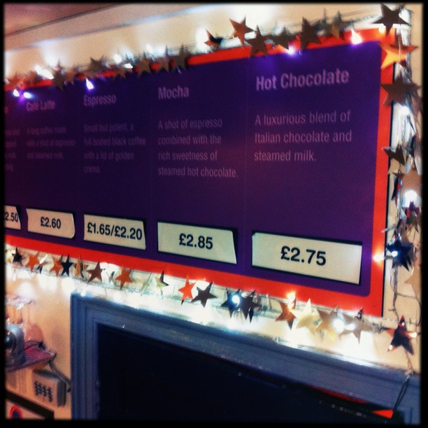 A coffee shop menu board displaying drink options with prices: Espresso £2.60, Mocha £1.65/£2.20, and Hot Chocolate £2.75, decorated with Christmas stars.