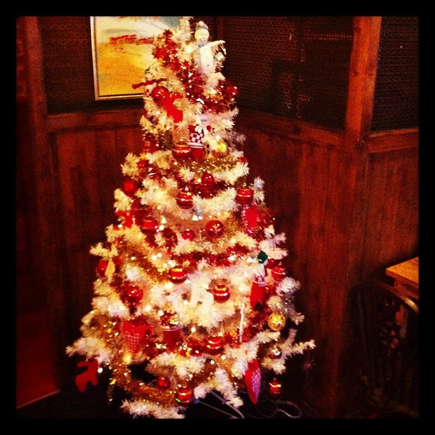 A white Christmas tree decorated with red and gold ornaments and lights, positioned in a room with wooden panelling.