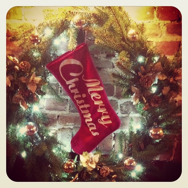 A red Christmas stocking with gold lettering hangs on a decorated wreath with lights and ornaments.