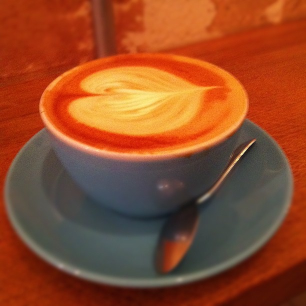 A cappuccino with latte art sits on a light blue saucer with a spoon, on a warm orange surface.