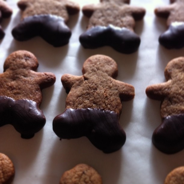 Gingerbread men biscuits with dark chocolate coating on their lower halves, arranged on parchment paper.