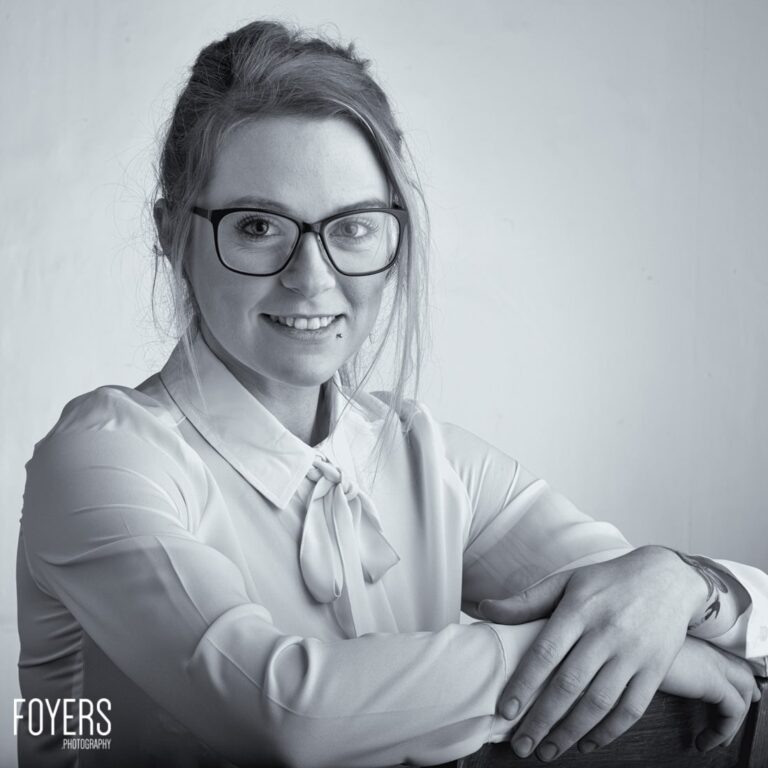 A woman wearing glasses and a white blouse with a tied collar smiles at the camera against a plain light background.