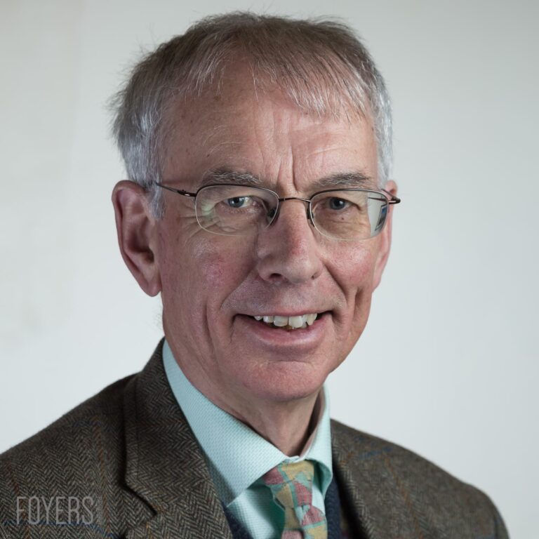 A man with grey hair and glasses wearing a brown tweed jacket and patterned tie, smiling at the camera against a white background.