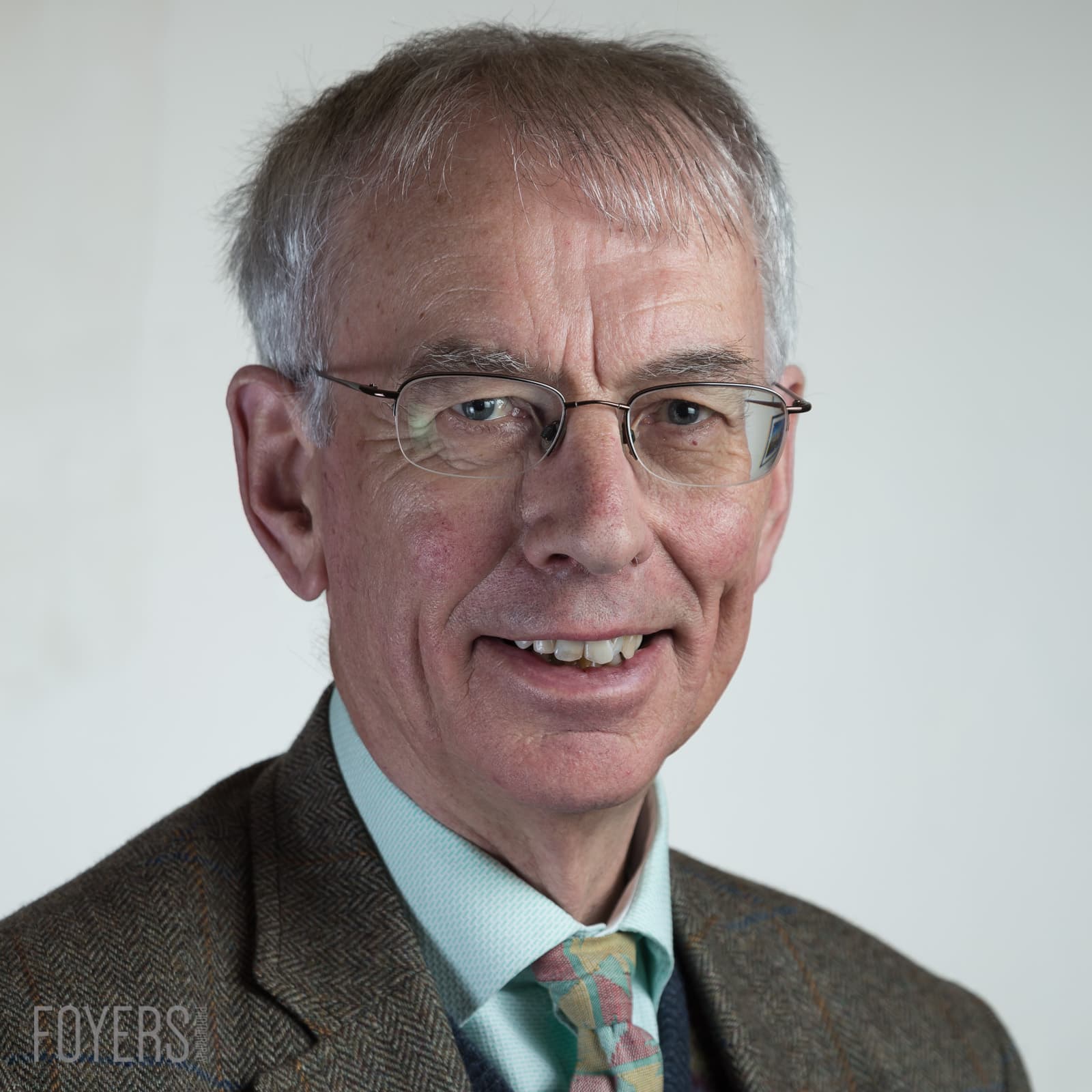 A man with grey hair and glasses wearing a brown tweed jacket and patterned tie, smiling at the camera against a white background.