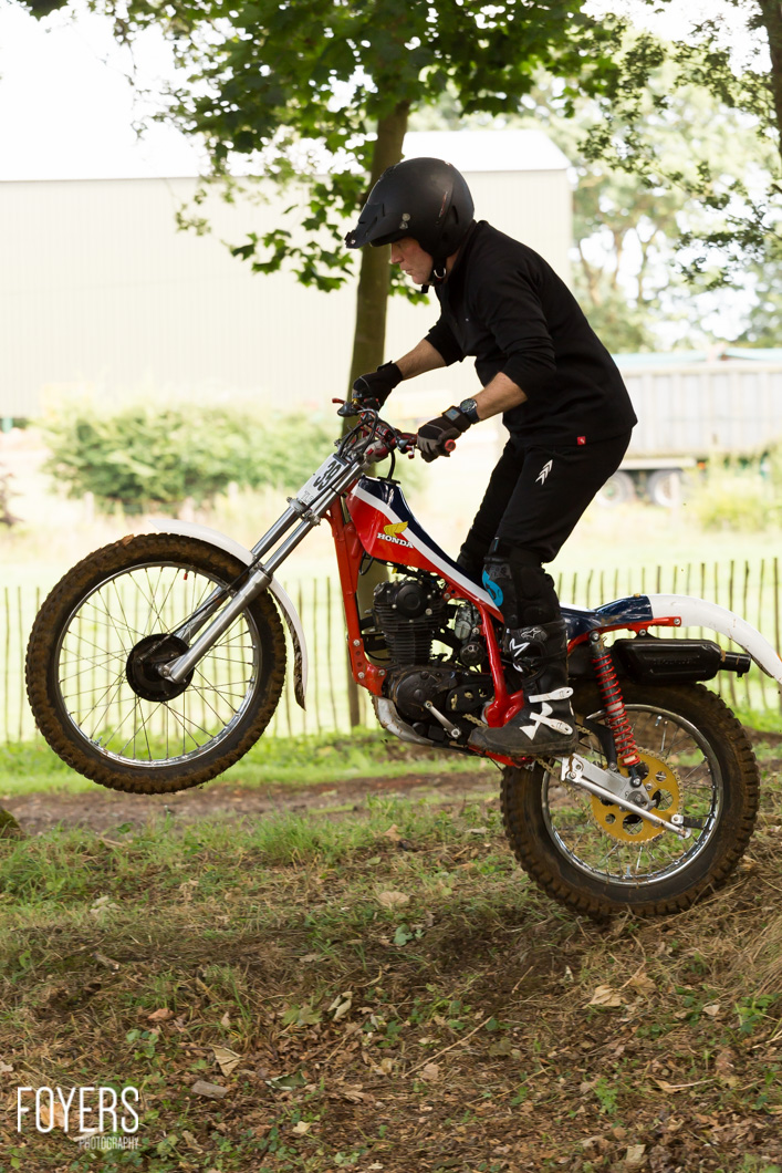 A person in black clothing and helmet performs a wheelie on a red Honda trials motorcycle on dirt terrain with grass and trees in background