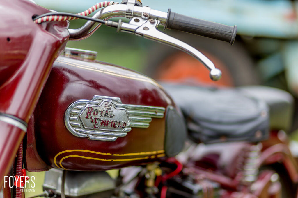 Close-up of a Royal Enfield motorcycle fuel tank with chrome badge and handlebar, showing maroon paintwork with yellow pinstriping