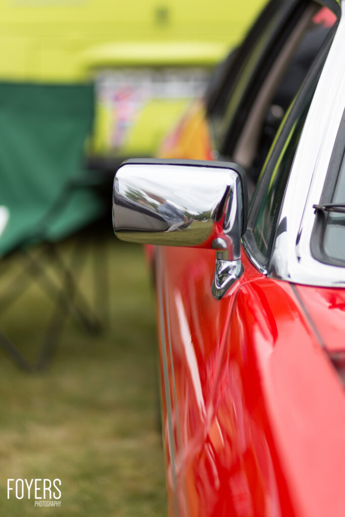 Chrome wing mirror on red and white classic car with blurred yellow and green background