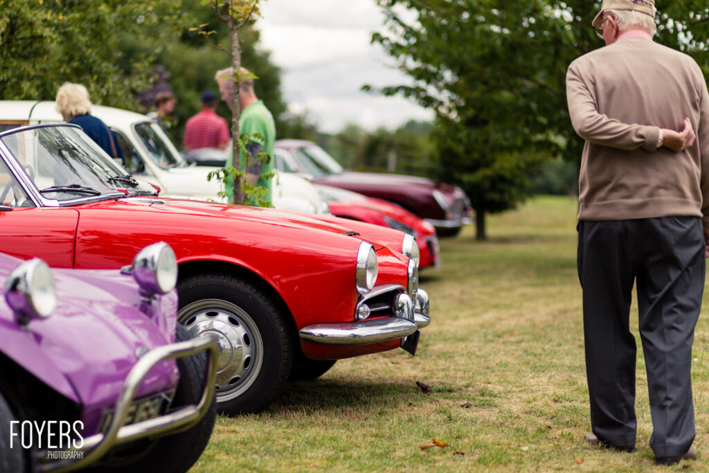 A man in beige jacket views classic cars lined up on grass at a car show, including a red and purple vintage vehicle in the foreground.