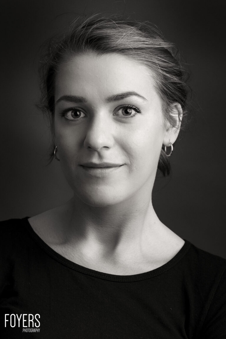 A person with short hair swept back, wearing hoop earrings and a black top, looking directly at camera in black and white portrait
