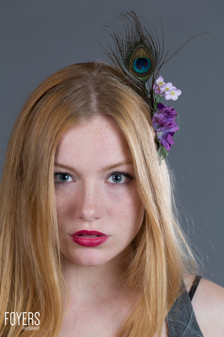Young woman with long blonde hair wearing peacock feather and purple flowers as hair accessory, looking at camera
