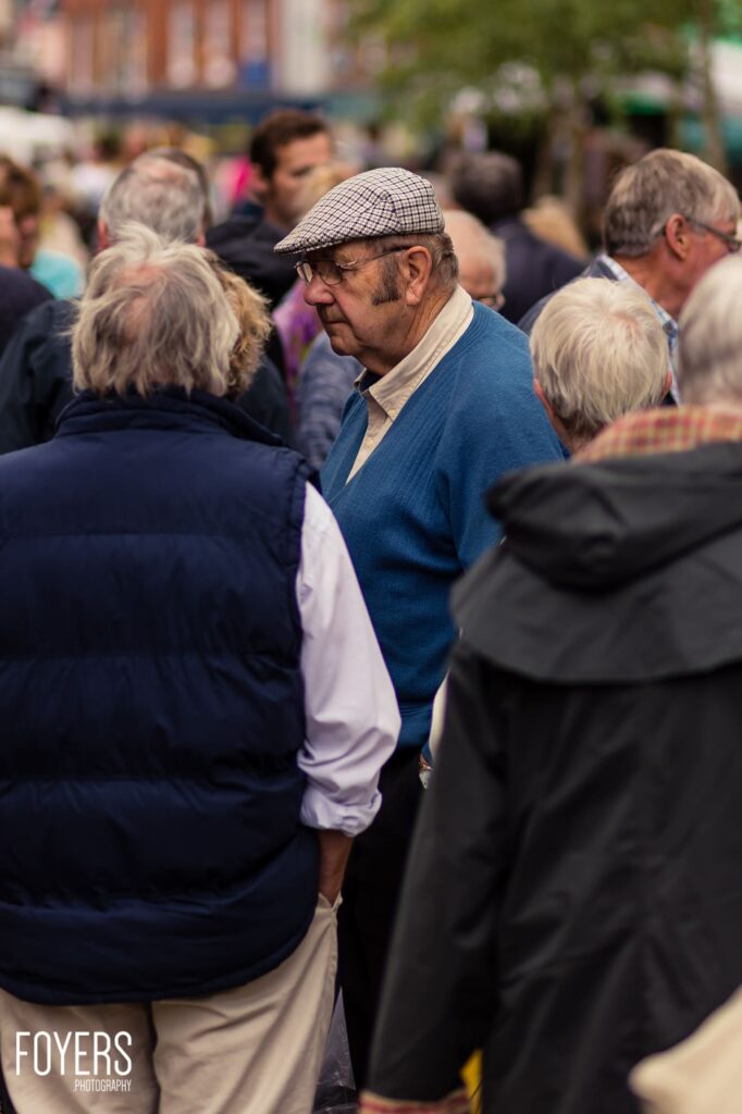 An elderly man wearing a flat cap, glasses and blue sweater stands among a crowd of people at an outdoor gathering
