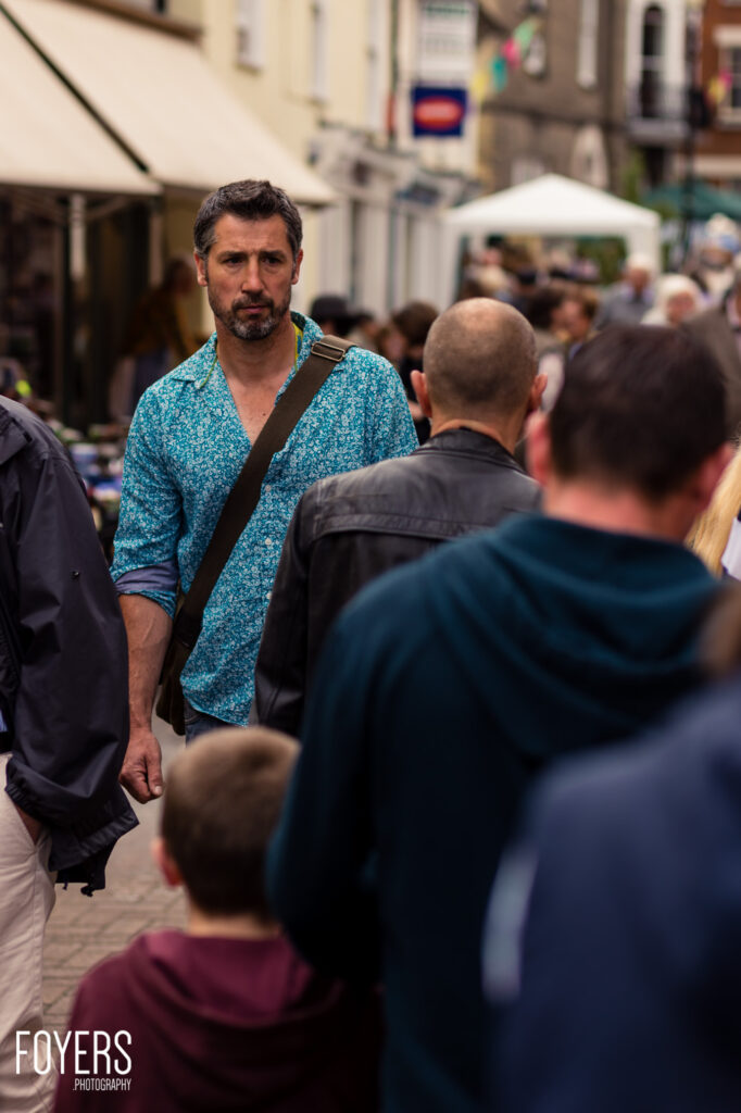 A man in a turquoise floral shirt with a shoulder bag walks through a crowded pedestrian street looking directly at the camera.