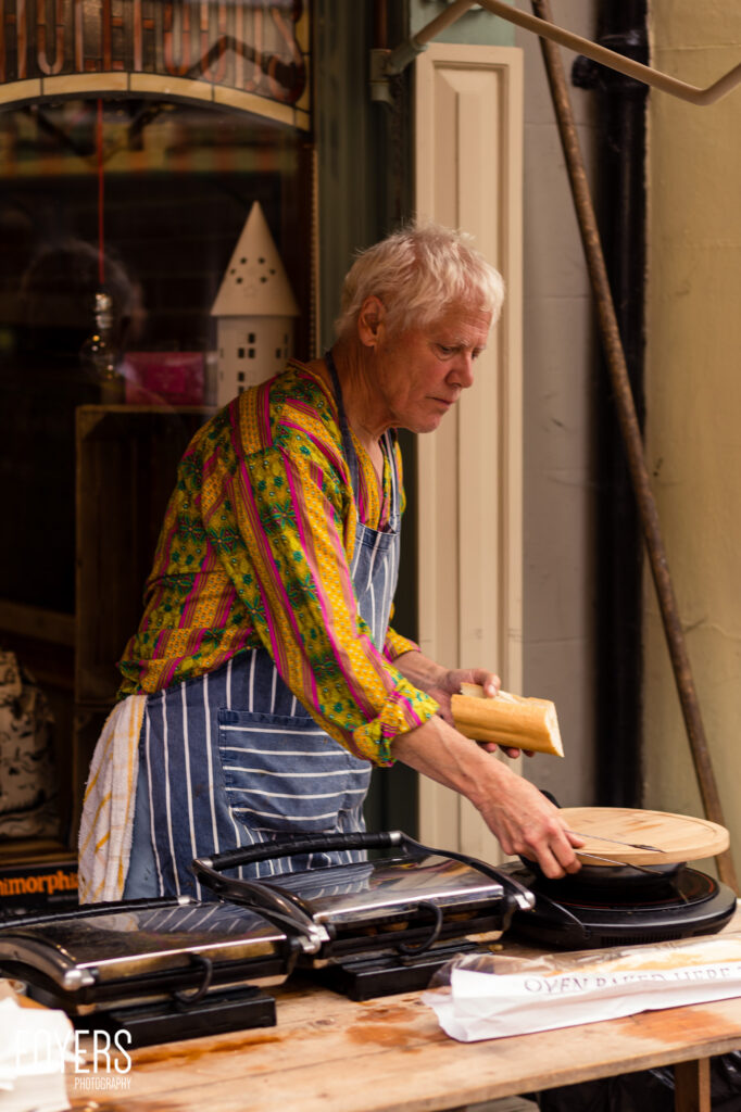 An older person with white hair wearing a colourful patterned jacket and striped apron prepares food at a counter with cooking equipment.