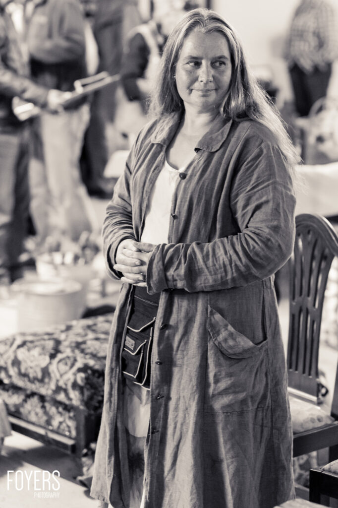 A woman with long hair wearing a button-up coat dress stands indoors with blurred figures visible in the background
