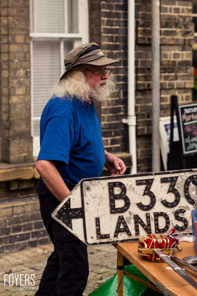 A bearded man in a blue shirt and cap holds a vintage B3301 Lands road sign at an outdoor market stall