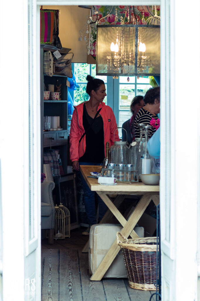 A woman in a red jacket and black top stands in a cottage-style dining room with wooden table and chandelier