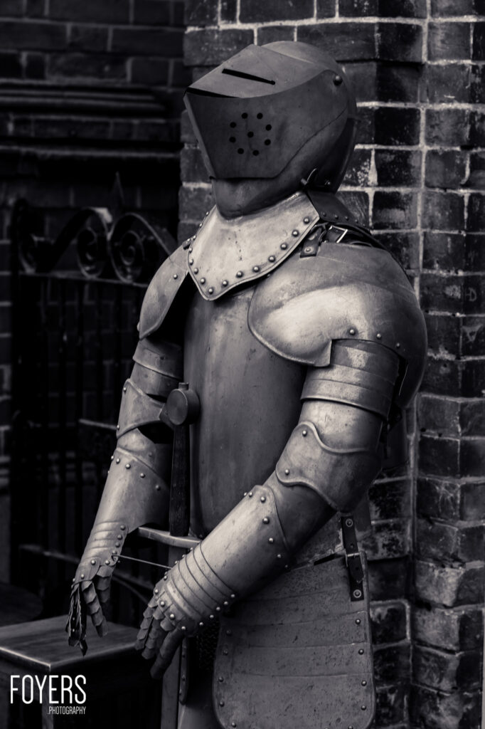 A suit of medieval plate armour standing against a brick wall, featuring a distinctive helmet with breathing holes and articulated metal plates covering the body and limbs.