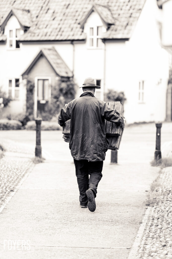 A person wearing a hat and jacket walks away from camera carrying a wooden box, with cottages in the background