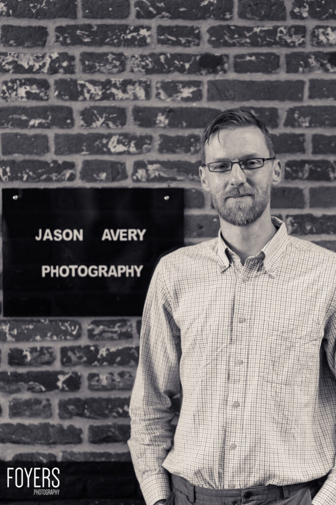 A man with glasses and a beard wearing a checked shirt stands in front of a brick wall with a photography business sign
