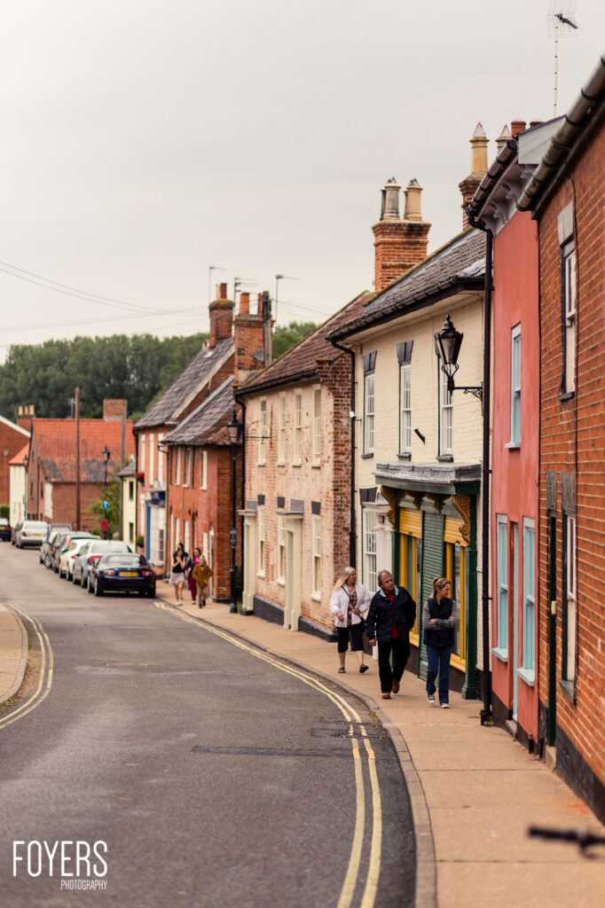 A quiet street in a historic English town with traditional brick buildings, chimneys, and pedestrians walking along the pavement.