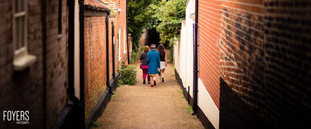 Three people walking away down a narrow brick-walled path between buildings, one wearing a blue coat and pink trousers