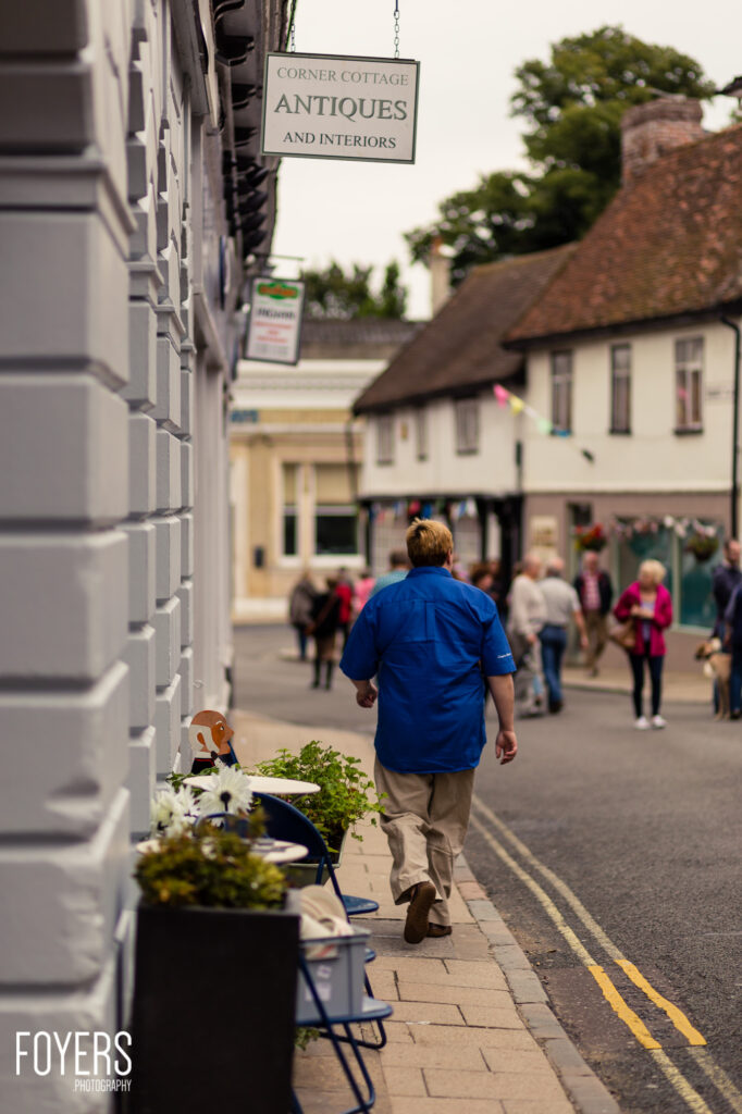 A person in a blue shirt walks along a pavement past planters and shop fronts on a busy high street with historic buildings