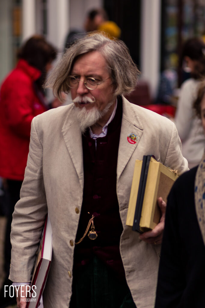 An older man with grey hair and beard wearing glasses and a beige blazer carries books while walking through a crowded indoor space