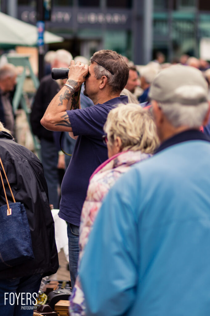 A man with a tattoo on his arm looks through binoculars in a crowd of people on a busy street.