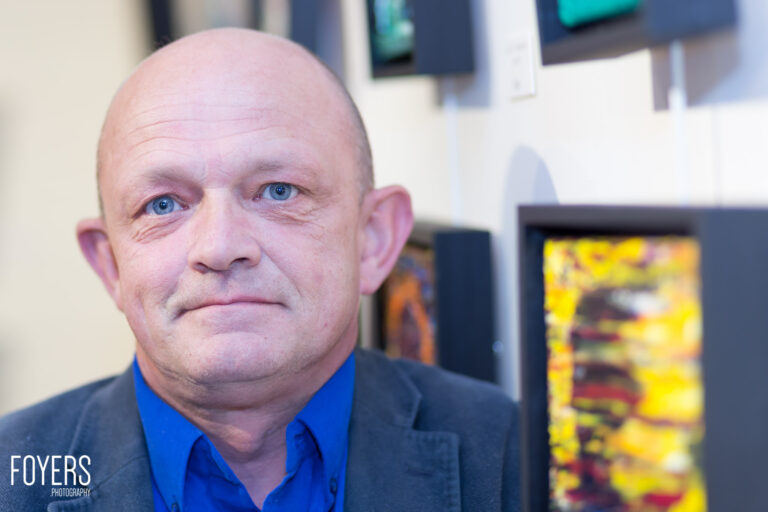 A bald man with blue eyes wearing a blue shirt and dark blazer looks directly at the camera in an office setting