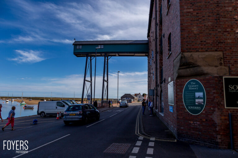 A coastal road passes under an elevated grain storage building on stilts, with boats moored in shallow water and brick buildings nearby