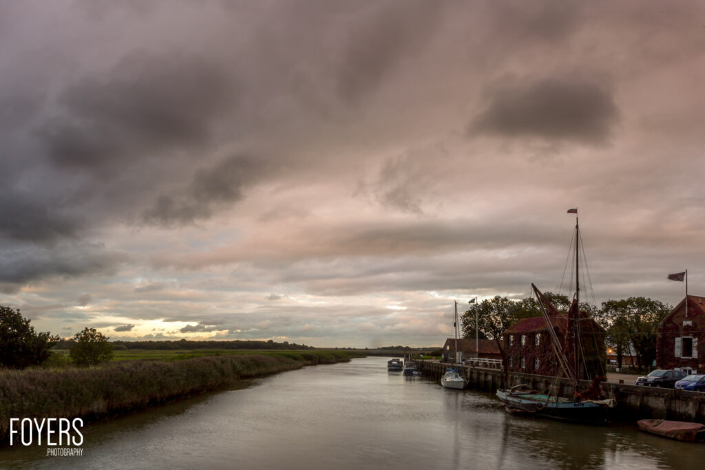 A moody river scene with boats moored alongside brick buildings under dramatic cloudy skies, with flat marshland visible