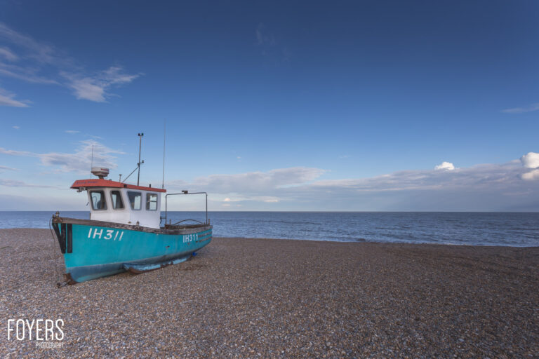 A turquoise fishing boat with registration IH311 rests on a pebble beach under a blue sky with wispy clouds