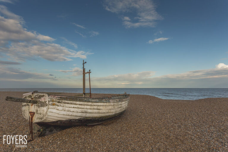 An old weathered white rowing boat sits on a pebble beach with the sea and cloudy sky in the background