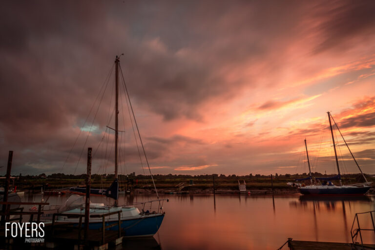 Sailboats moored in a harbour at sunset with dramatic orange and pink clouds reflected in calm water