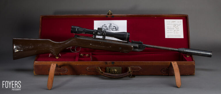 A rifle with wooden stock and telescopic sight displayed in an open brown leather case lined with red velvet, accompanied by documentation.