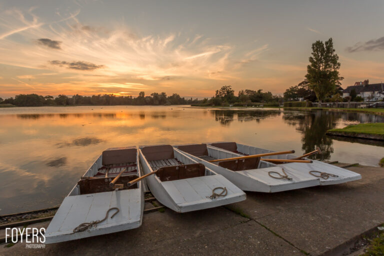Three rowing boats beached on concrete beside a calm river at sunset, with trees and houses reflected in the water.