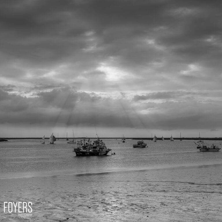 Moored fishing boats on shallow water with beach in foreground and dramatic cloudy sky above. Black and white photograph.