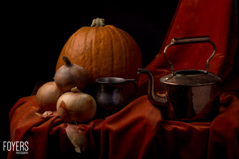 A still life arrangement featuring a pumpkin, onions, dark metal vessels and red fabric against a black background.