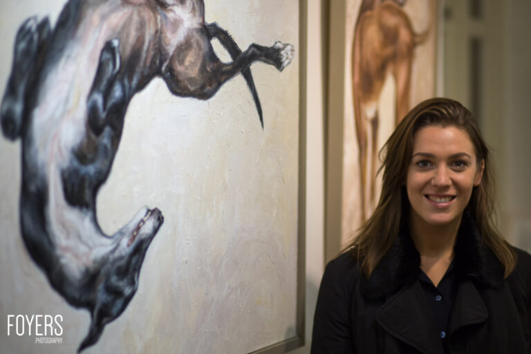 Woman smiling at camera beside mural of primate holding a needle, gallery or museum setting.