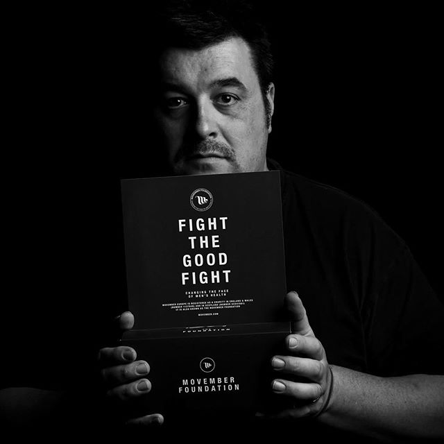 A man in dark clothing holds a black box printed with FIGHT THE GOOD FIGHT and the Movember Foundation logo against a black background.