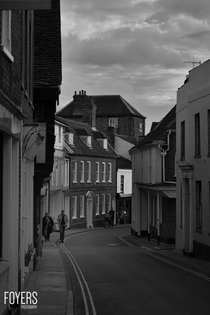 A narrow high street lined with Georgian and Victorian buildings in black and white. A few pedestrians walk along the pavement under cloudy skies.