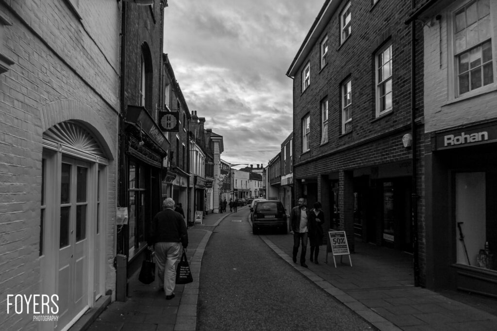 A narrow high street lined with brick buildings. A few pedestrians walk past shops. A car is parked in the centre. Cloudy sky overhead.