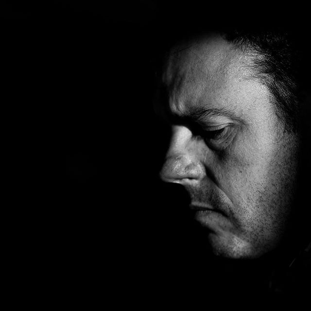A man's face in profile, shown in black and white. He has his eyes closed and appears contemplative against a dark background.